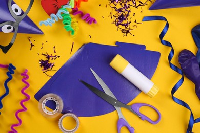 Photo of Handmade party hats, template and tools on yellow background, flat lay