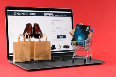 Photo of Online store. Laptop, small shopping cart, purchases and credit card on red background