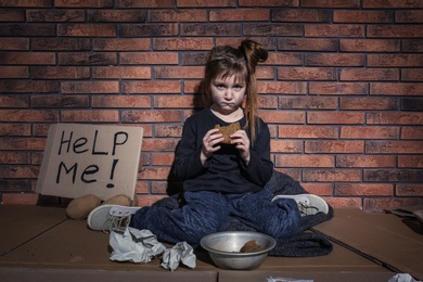 Poor little girl with bread and HELP ME sign on floor near brick wall