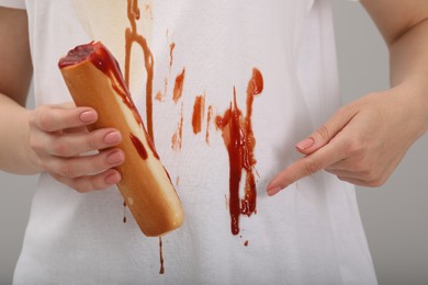 Woman holding hotdog and showing stain from sauce on her shirt against light grey background, closeup