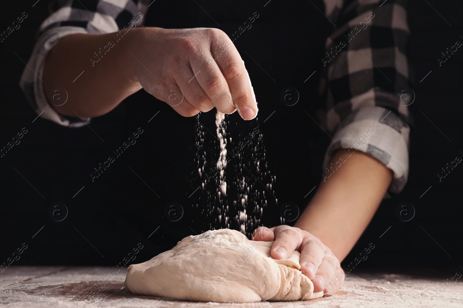 Photo of Man sprinkling flour over dough at wooden table on dark background, closeup