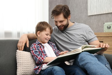 Photo of Happy dad and son reading book together on sofa at home