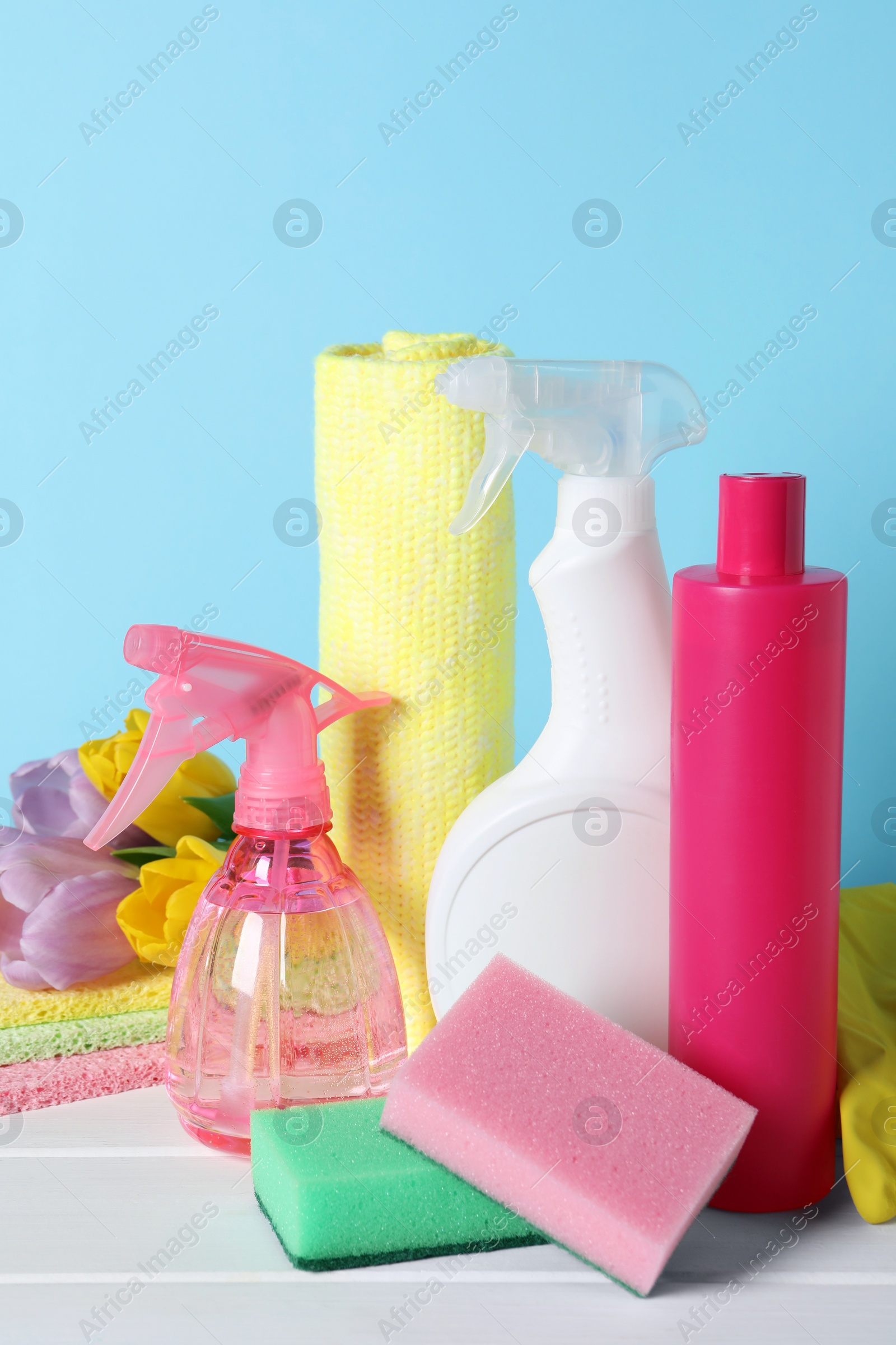 Photo of Spring cleaning. Detergents, flowers and sponges on white wooden table against light blue background