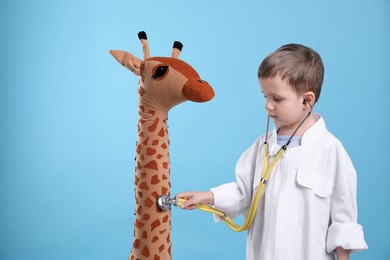 Photo of Cute little boy in pediatrician's uniform playing with stethoscope and toy giraffe on light blue background