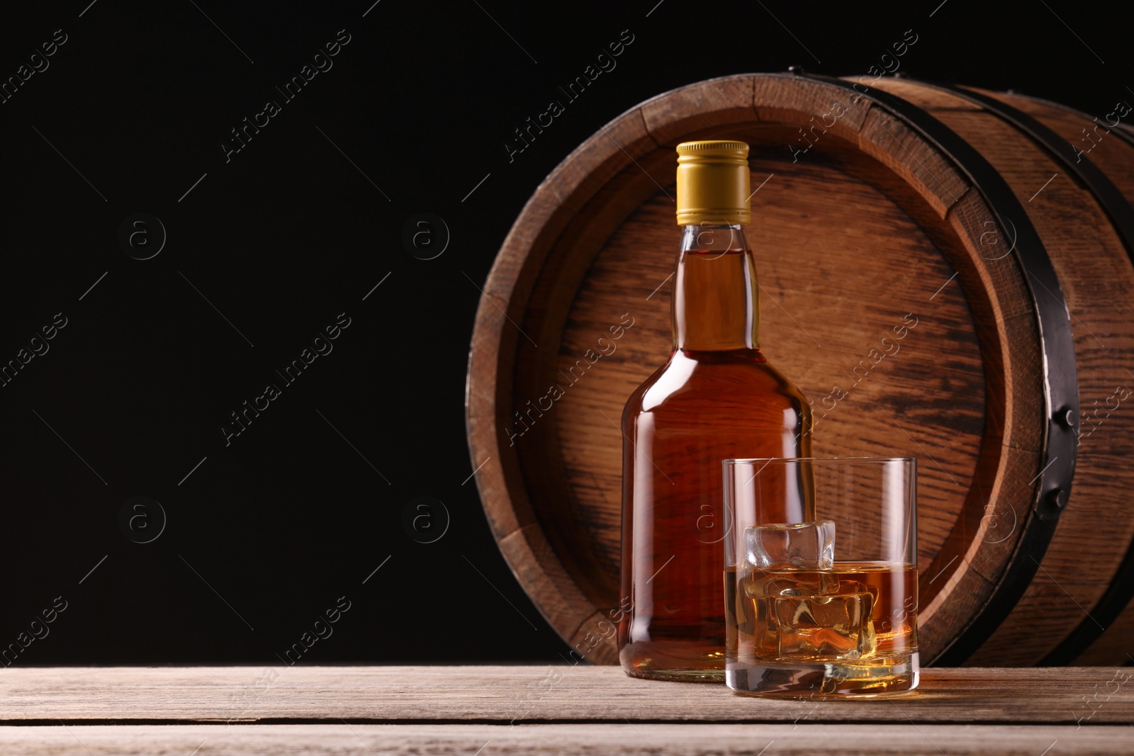 Photo of Whiskey with ice cubes in glass, bottle and barrel on wooden table against black background, space for text