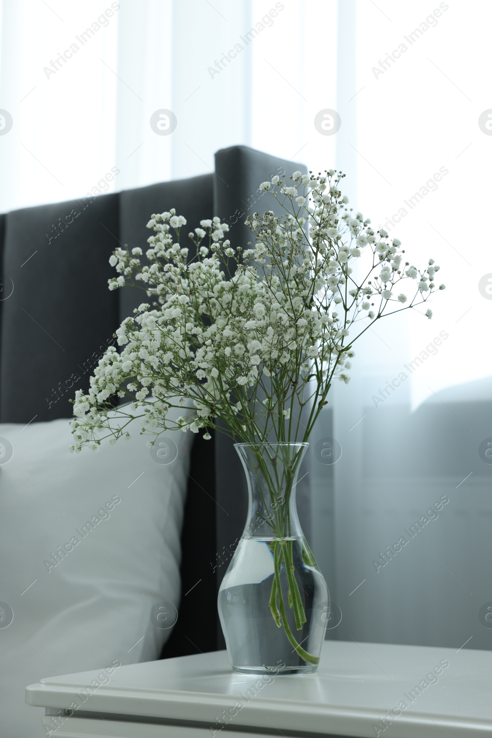 Photo of Beautiful gypsophila flowers in glass vase on white bedside table indoors