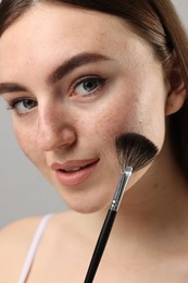 Beautiful woman with freckles applying makeup with brush on grey background, closeup