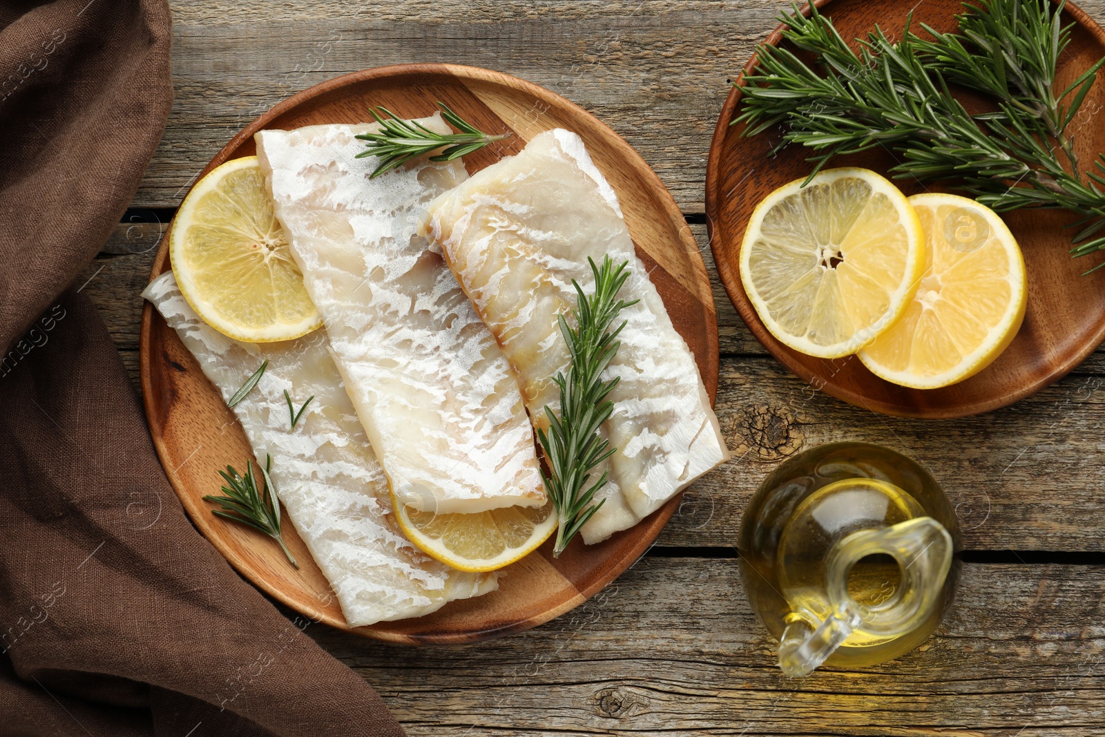 Photo of Fresh raw cod fillets with rosemary, lemon and oil on wooden table, flat lay