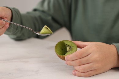 Photo of Boy eating tasty fresh kiwi with spoon at white marble table indoors, closeup