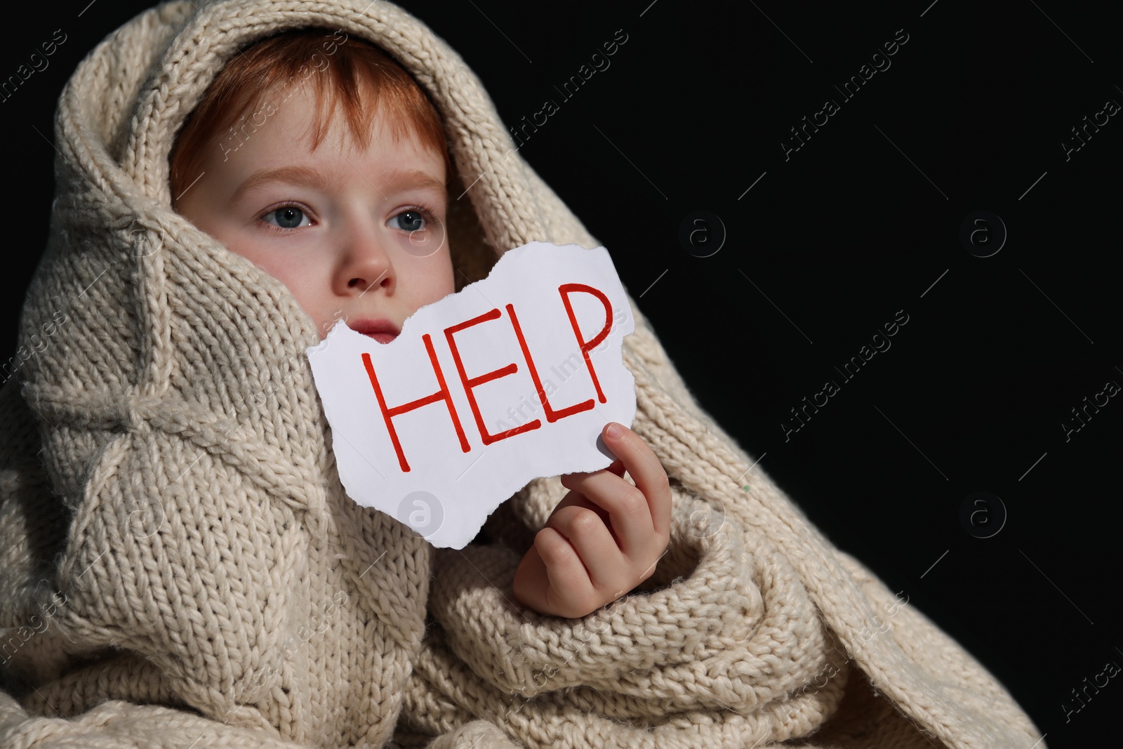 Photo of Scared little boy wrapped in blanket holding piece of paper with word Help on black background. Domestic violence concept