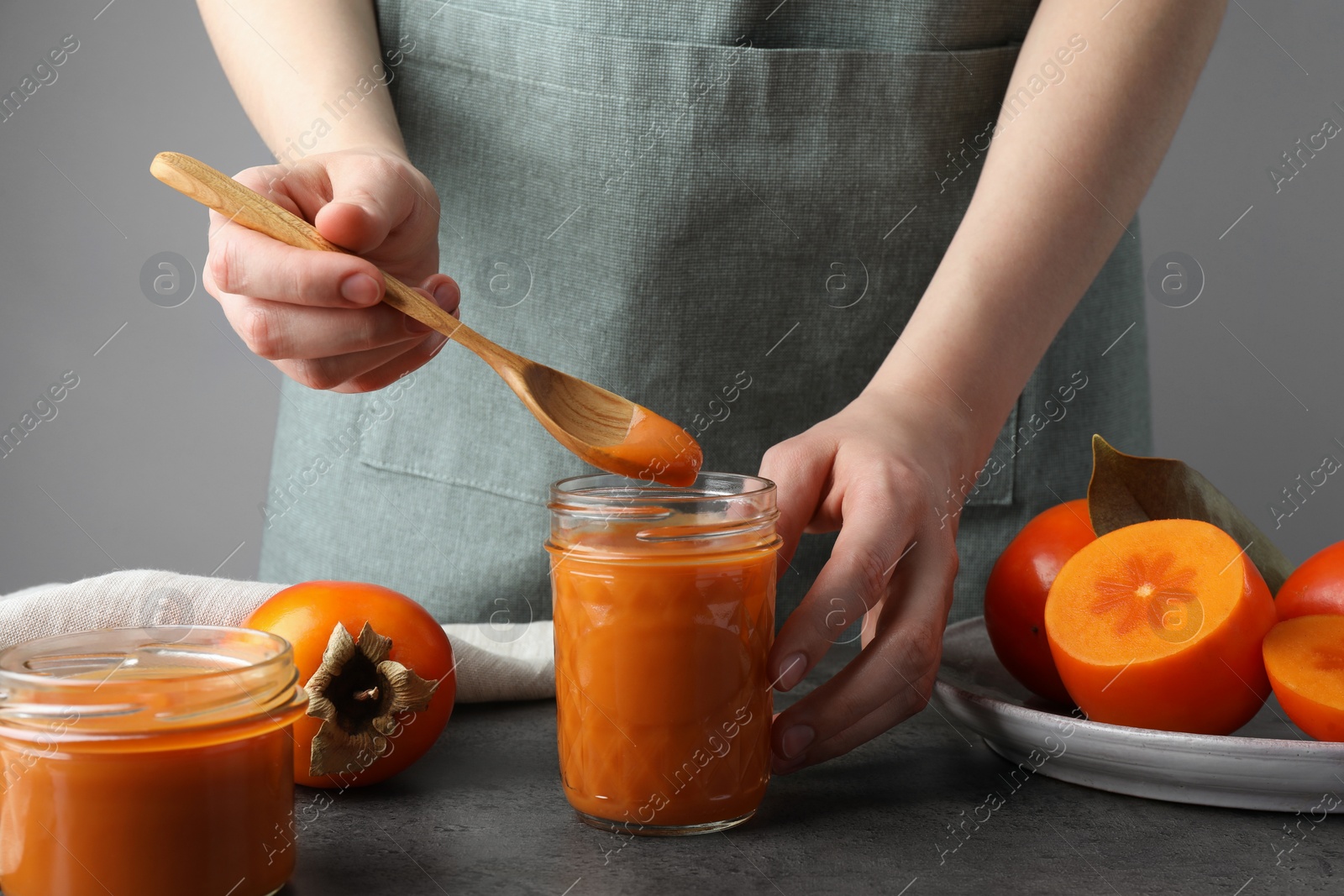 Photo of Woman taking delicious persimmon jam with spoon from glass jar at gray table, closeup