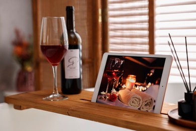 Photo of Wooden tray with tablet, glass of wine and bottle on bathtub in bathroom
