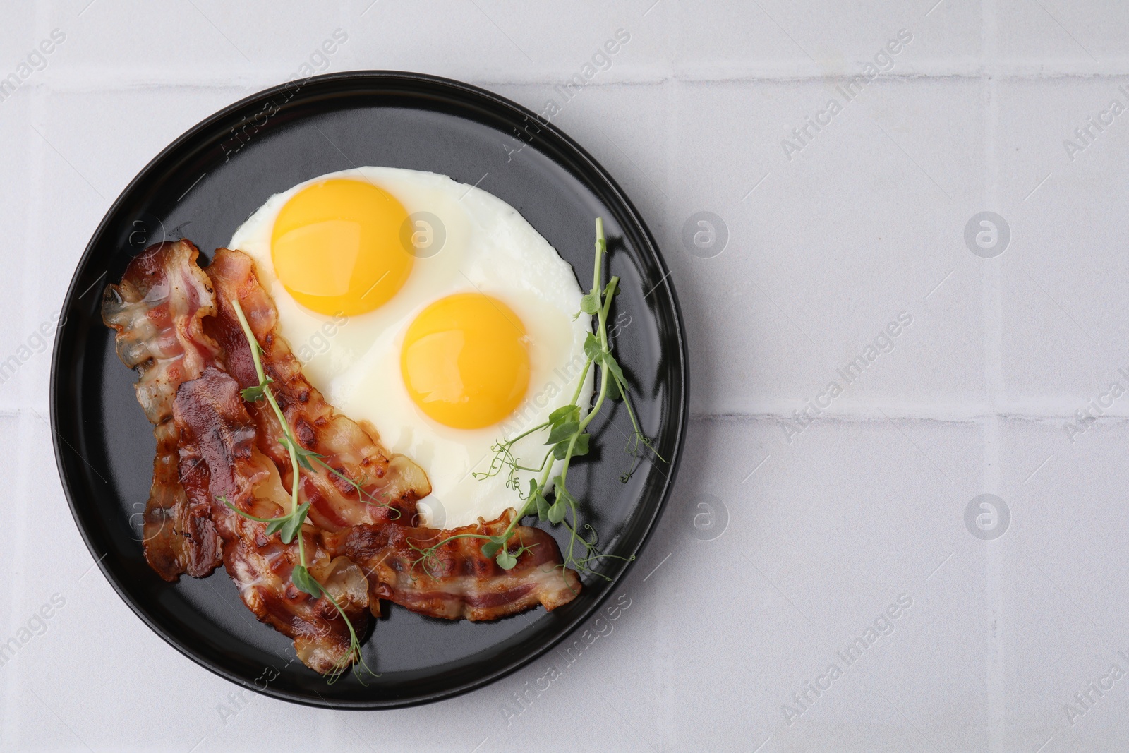 Photo of Fried eggs, bacon and microgreens on white tiled table, top view. Space for text