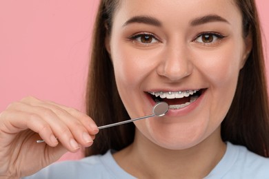 Smiling woman with braces holding dental mirror on pink background, closeup