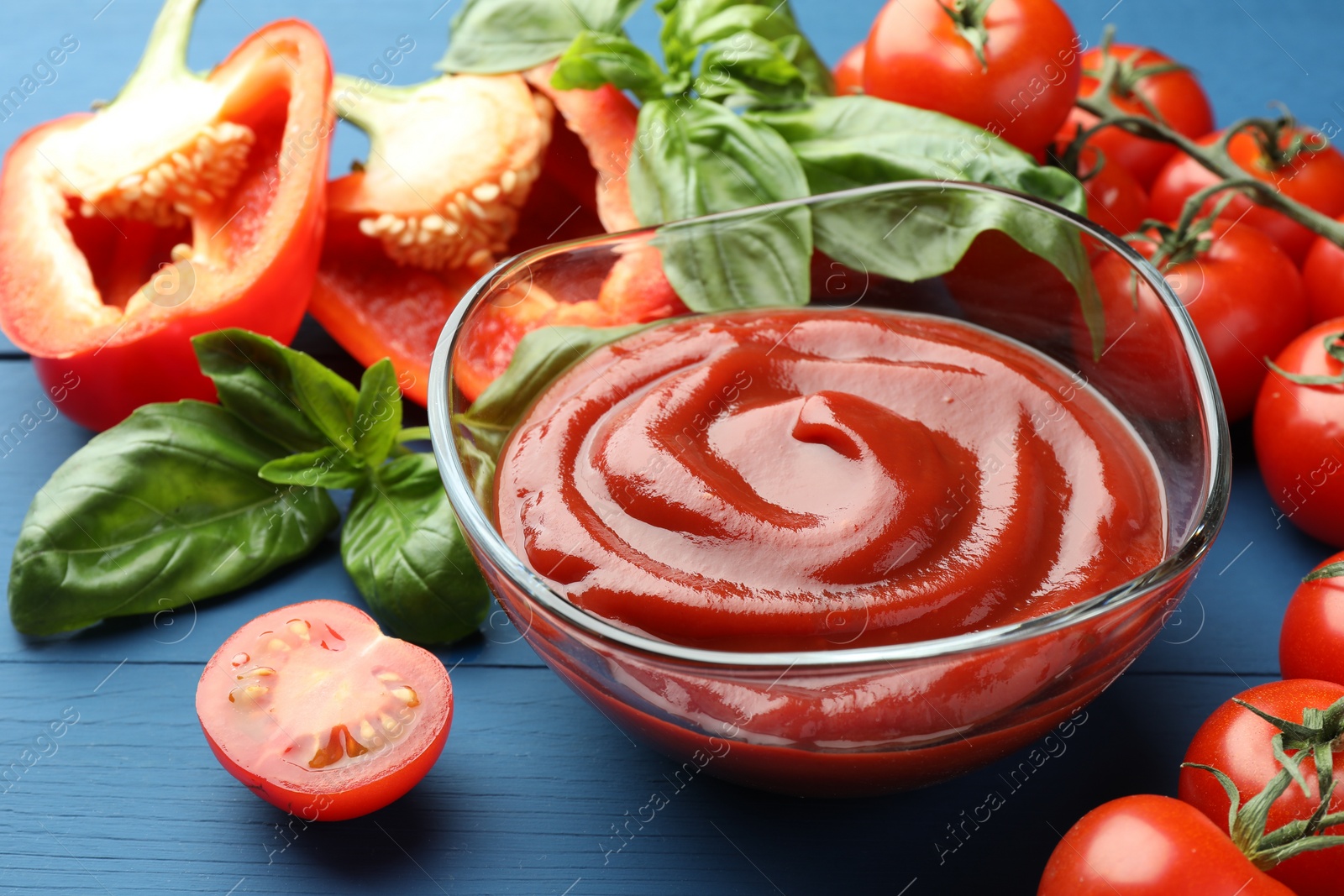 Photo of Bowl of tasty ketchup and ingredients on blue wooden table, closeup