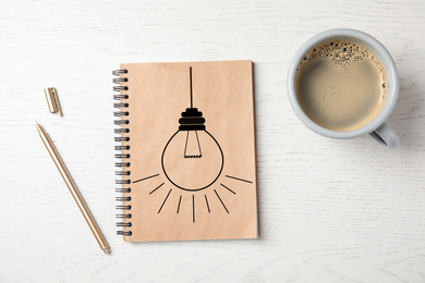 Image of Notebook with drawn lamp and cup of coffee on white wooden table, flat lay. New idea concept