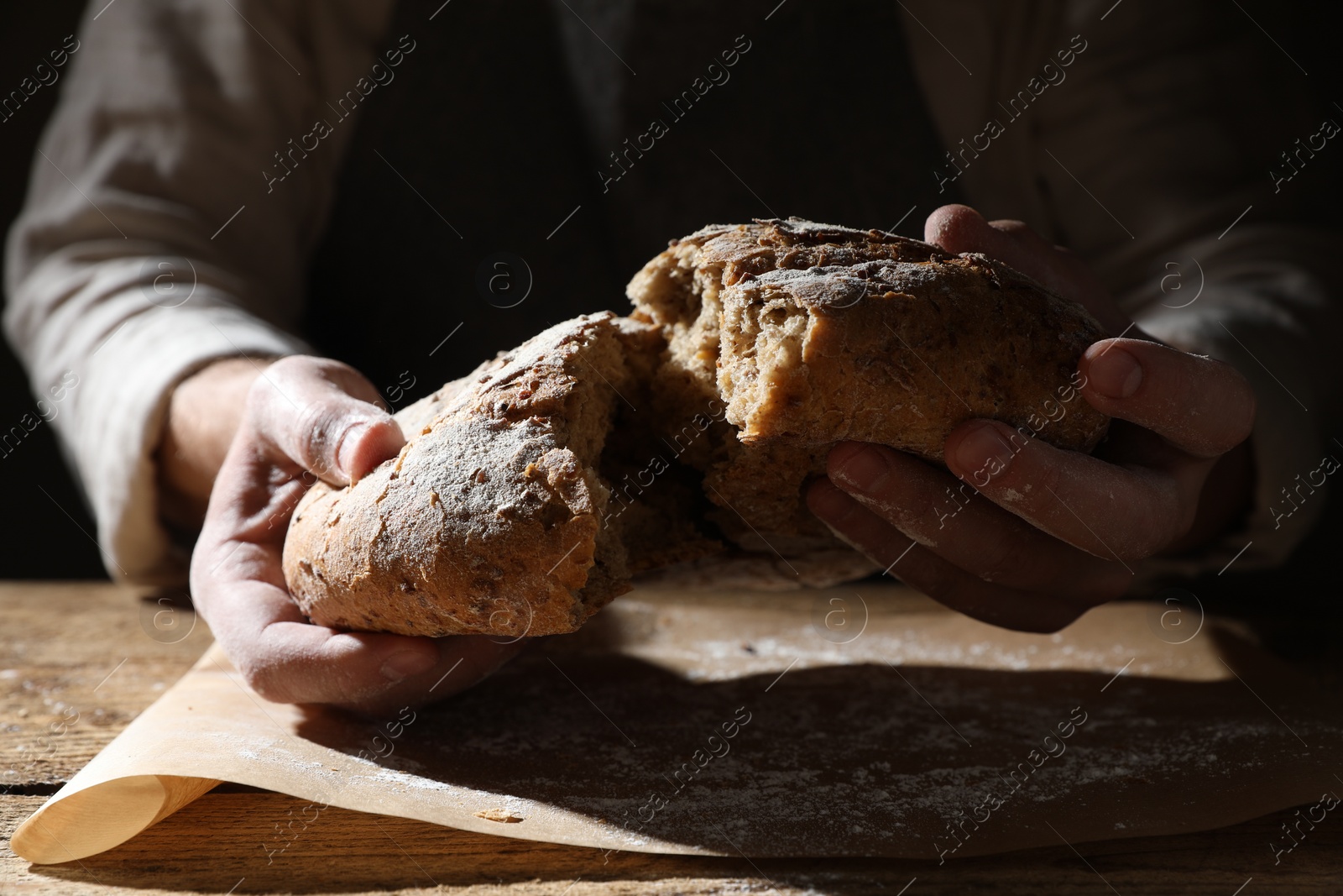 Photo of Man breaking loaf of fresh bread at wooden table, closeup