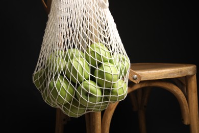 Green apples in net bag hanging on wooden chair against black background, closeup