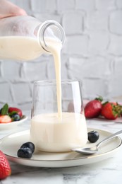 Woman pouring tasty yogurt into glass at white marble table, closeup