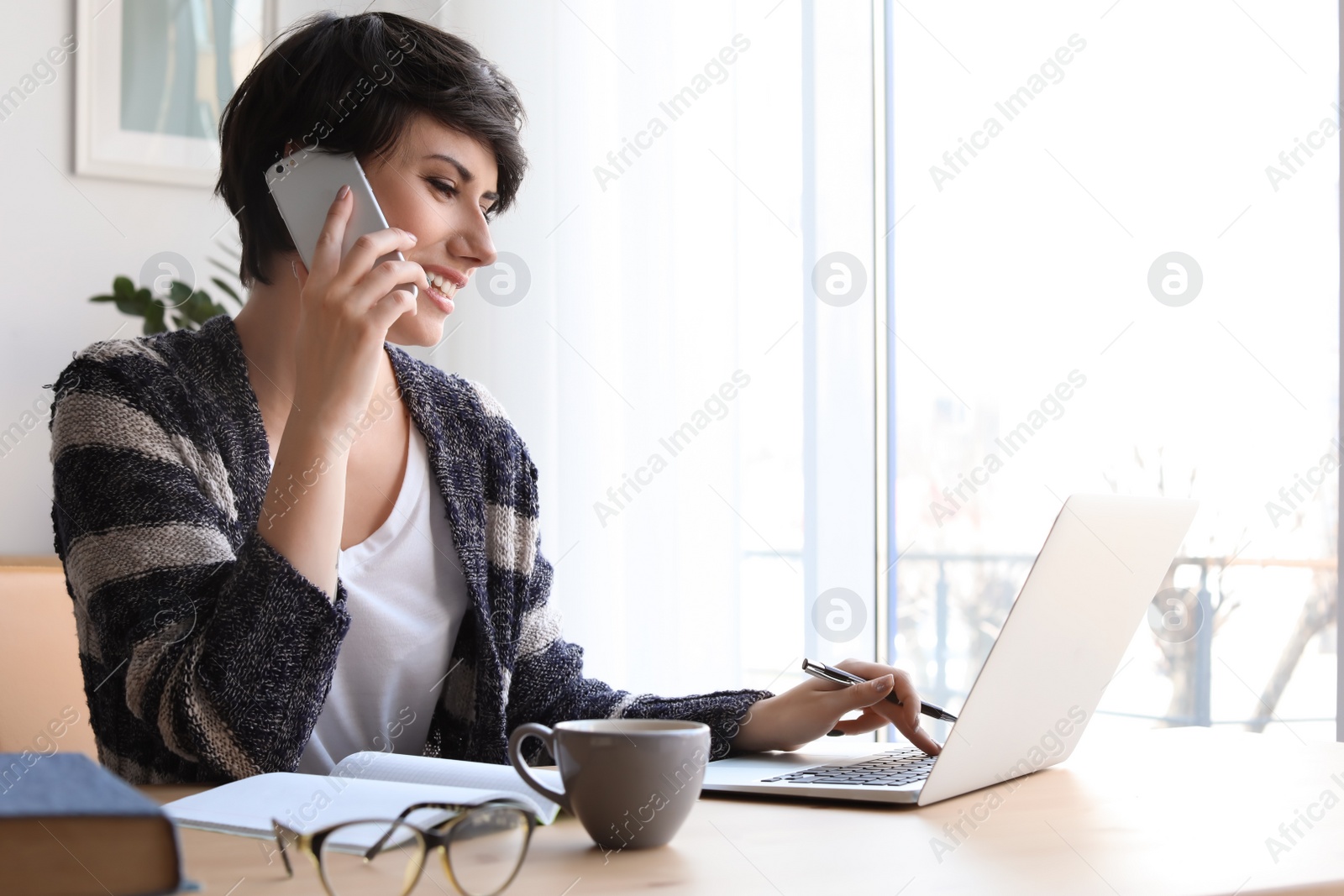 Photo of Young woman talking on mobile phone while working with laptop at desk. Home office