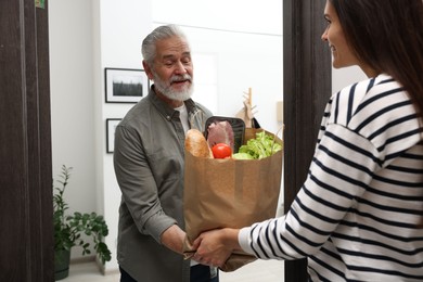 Courier giving paper bag with food products to senior man indoors