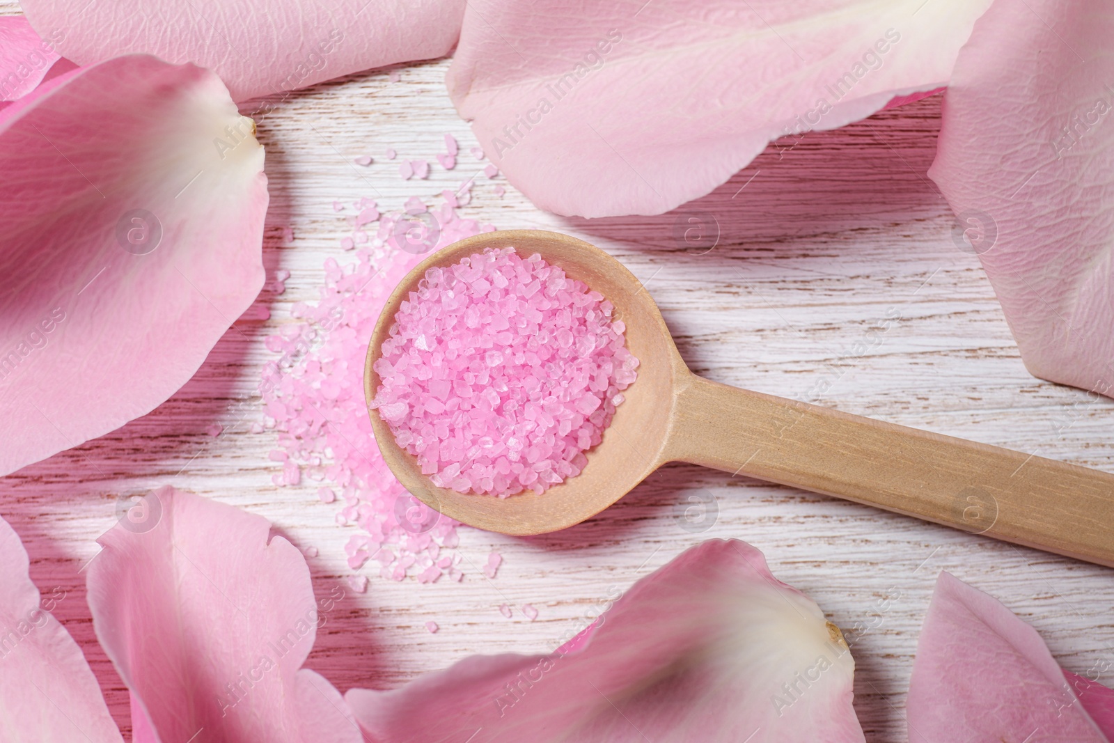Photo of Spoon with pink sea salt and petals of roses on white wooden table, flat lay