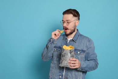 Handsome young man eating tasty potato chips on light blue background