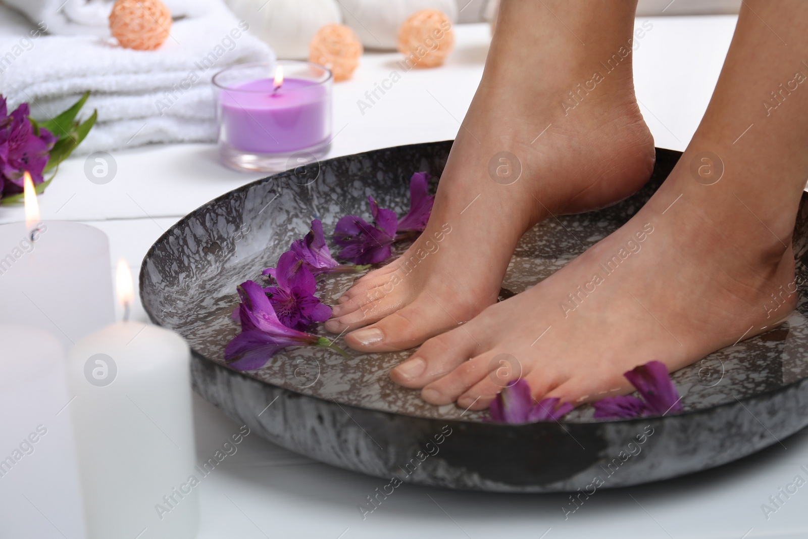 Photo of Woman soaking her feet in bowl with water and flowers on white floor, closeup. Spa treatment