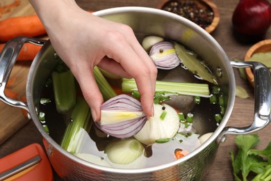 Cooking tasty bouillon. Woman putting onion into pot with different ingredients at wooden table, closeup