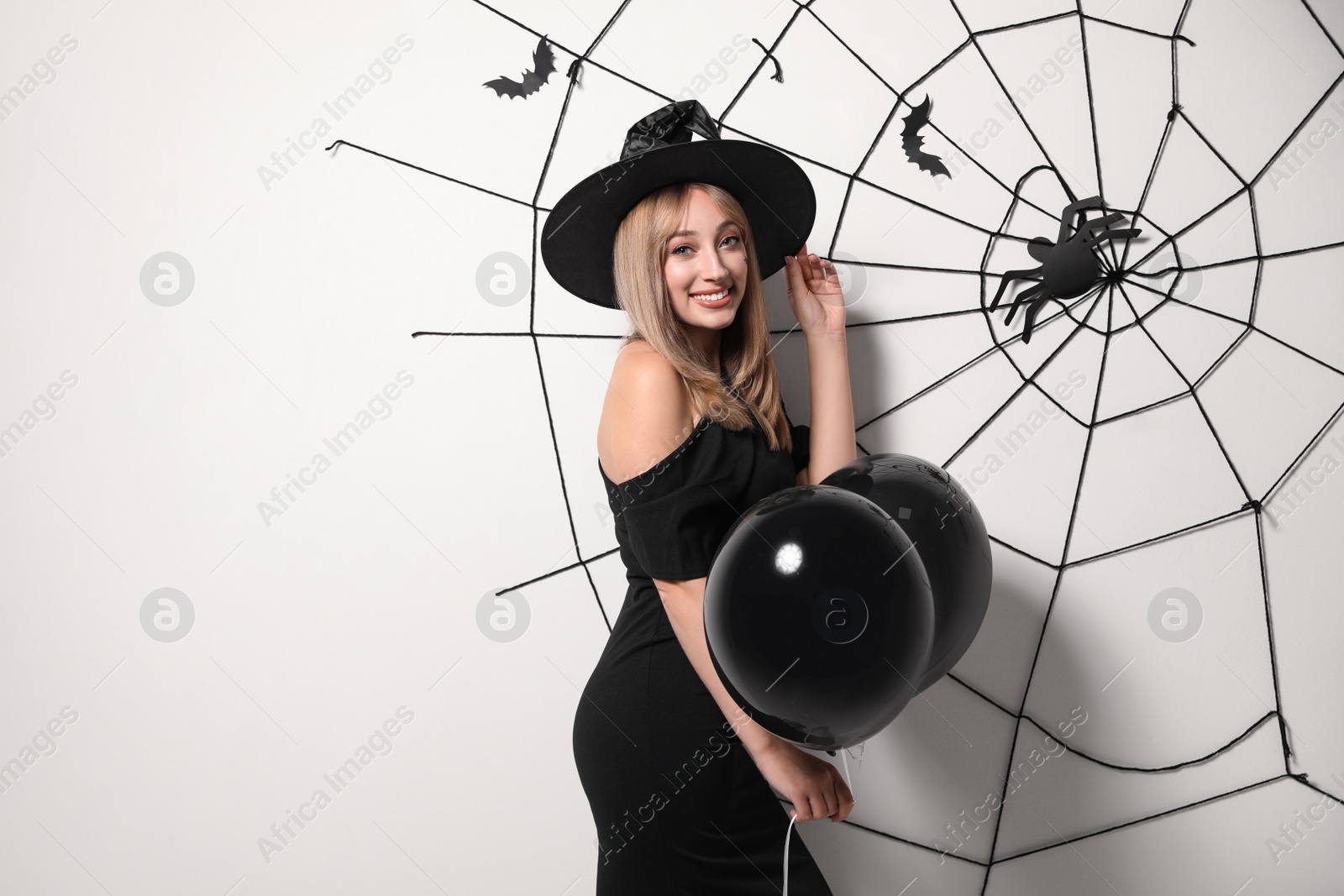 Photo of Woman in witch hat with balloons posing near white wall decorated for Halloween