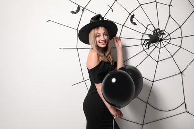 Woman in witch hat with balloons posing near white wall decorated for Halloween