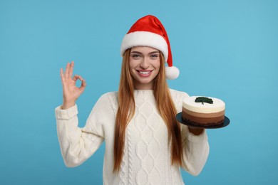 Photo of Young woman in Santa hat with tasty cake showing ok gesture on light blue background