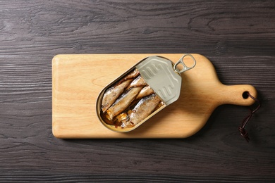 Board with tin can of sprats on wooden table, top view