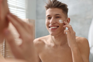 Photo of Handsome man applying moisturizing cream onto his face in bathroom
