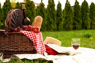 Photo of Picnic blanket with wicker basket, wine, food, sweater, straw hat, glass and book on green grass