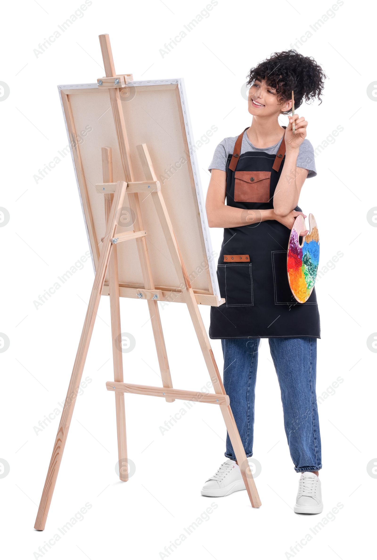 Photo of Young woman holding brush and artist`s palette near easel with canvas against white background