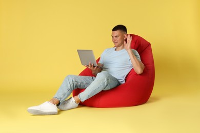 Photo of Handsome man with laptop waving hello on red bean bag chair against yellow background