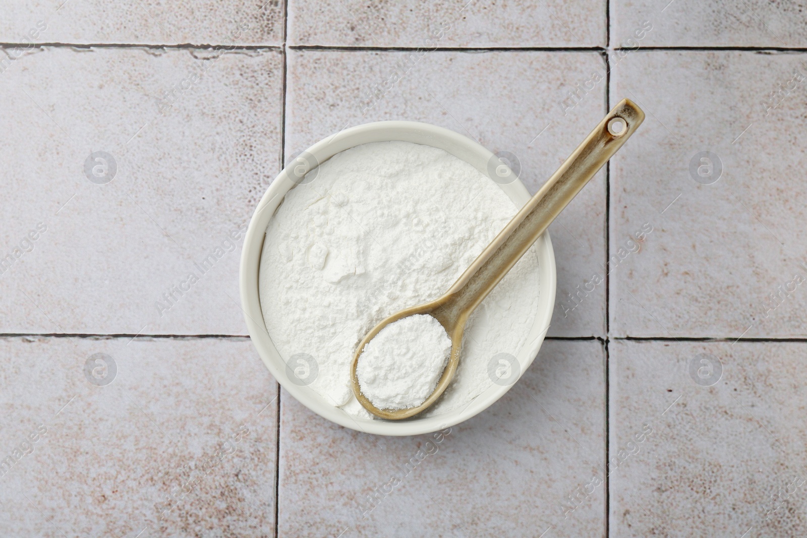 Photo of Baking powder in bowl and spoon on light tiled table, top view