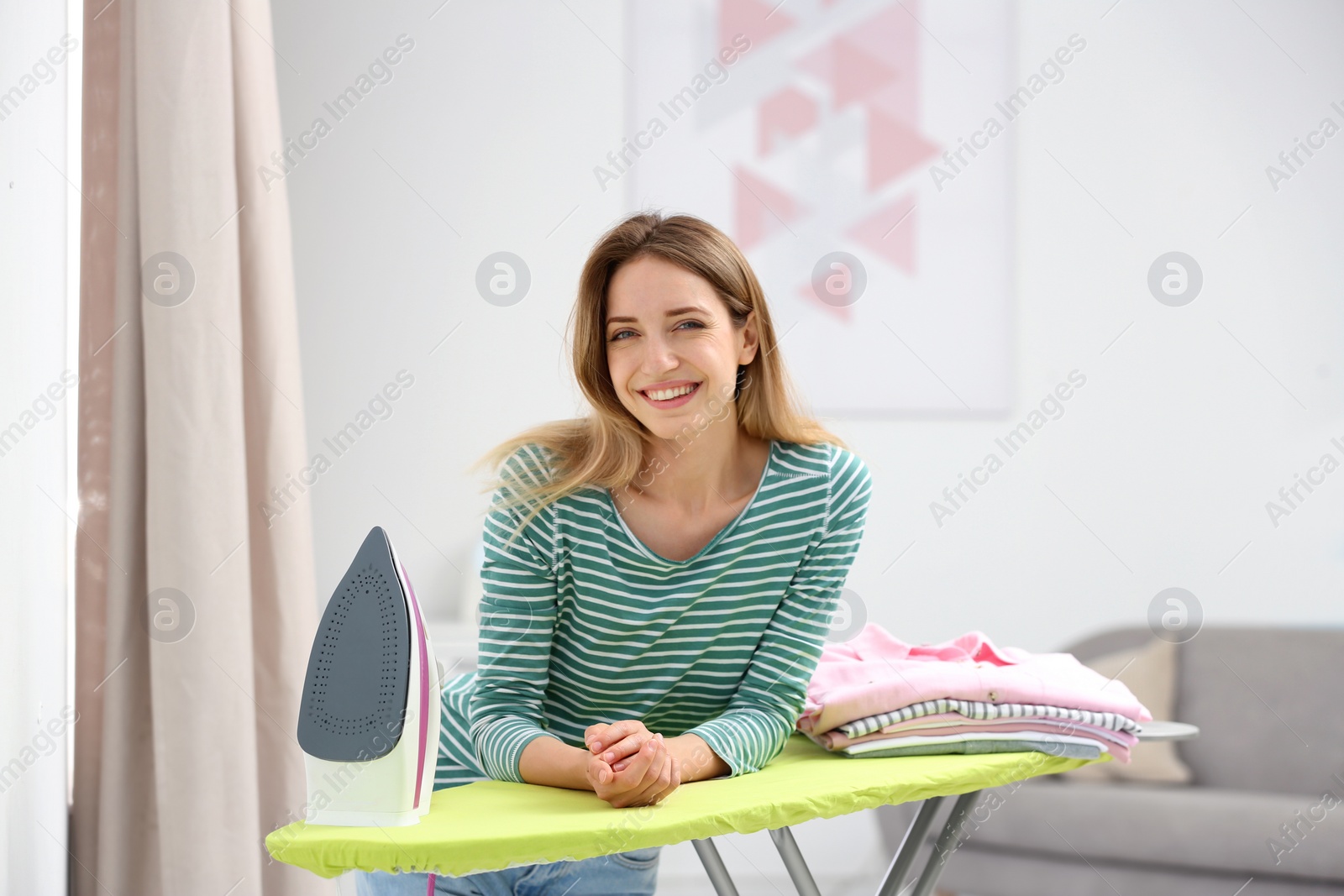 Photo of Young pretty woman with iron and clean laundry at board indoors