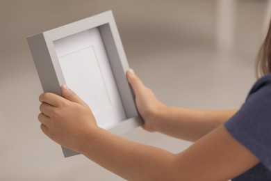 Photo of Little girl holding empty photo frame on blurred background, closeup