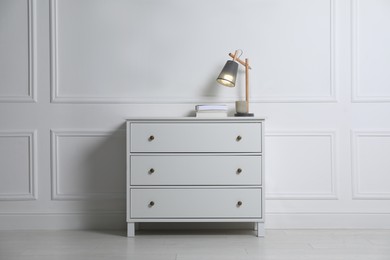 Photo of Lamp and books on chest of drawers near white wall indoors