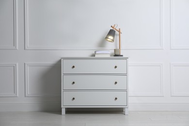 Lamp and books on chest of drawers near white wall indoors