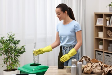 Photo of Garbage sorting. Smiling woman throwing glass bottle into trash bin in room