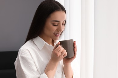 Photo of Beautiful woman with cup of drink near window at home, space for text. Lazy morning