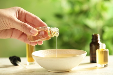 Photo of Woman pouring essential oil from glass bottle into bowl on table, closeup