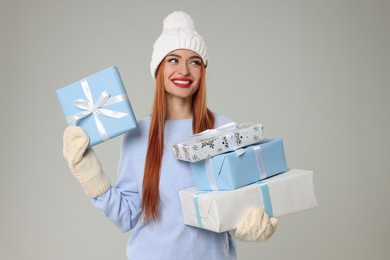 Young woman in hat and sweater with Christmas gifts on light grey background