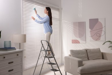 Photo of Woman on metal ladder wiping blinds at home