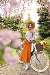 Beautiful young woman with bicycle and flowers in park on pleasant spring day