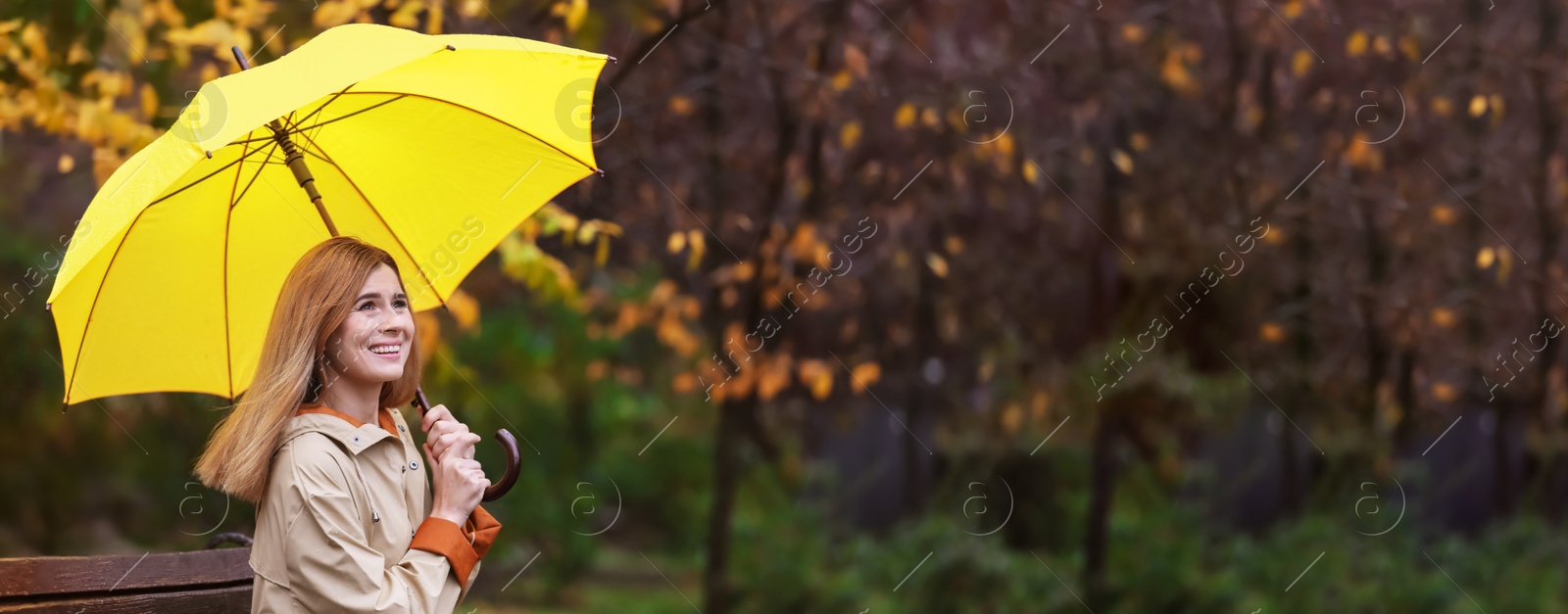 Image of Happy woman with umbrella sitting on bench in autumn park on rainy day, space for text. Banner design