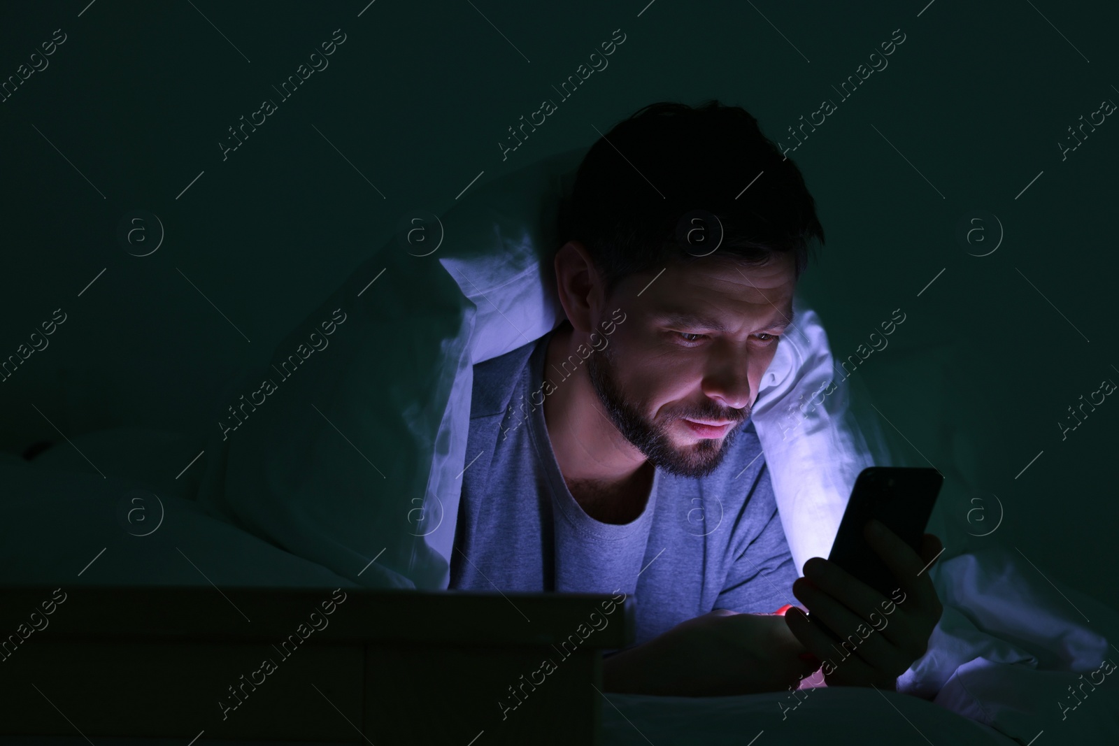 Photo of Man using smartphone under blanket in bed at night. Internet addiction